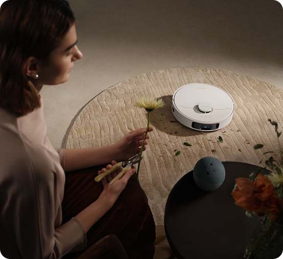 Kobieta przygotowuje bukiet kwiatów. Na stoliku znajduje się Amazon Alexa, a robot sprzątający odkurza dywan.